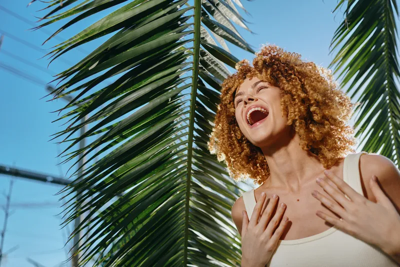 Laughing woman with curly hair enjoys sunny day outdoors near lush palm leaves, expressing happiness and natural beauty under clear blue sky.