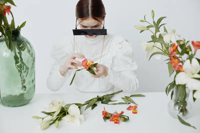 Stylish woman creating a floral arrangement, wearing a white blouse, with colorful flowers on a minimalist table, showcasing creativity and elegance