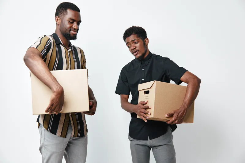 Two black men holding cardboard boxes for moving or delivery, standing on white background. African ethnicity, casual clothes, teamwork and cooperation in logistics or relocation.