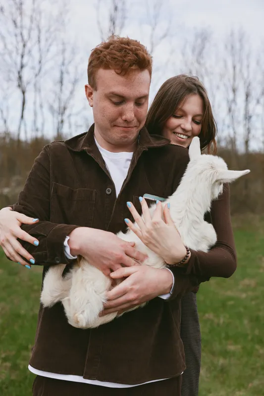 Happy couple outdoors in a green field cradle a white goat kid, smiling and affectionate, enjoying a rustic moment, casual clothing, natural light