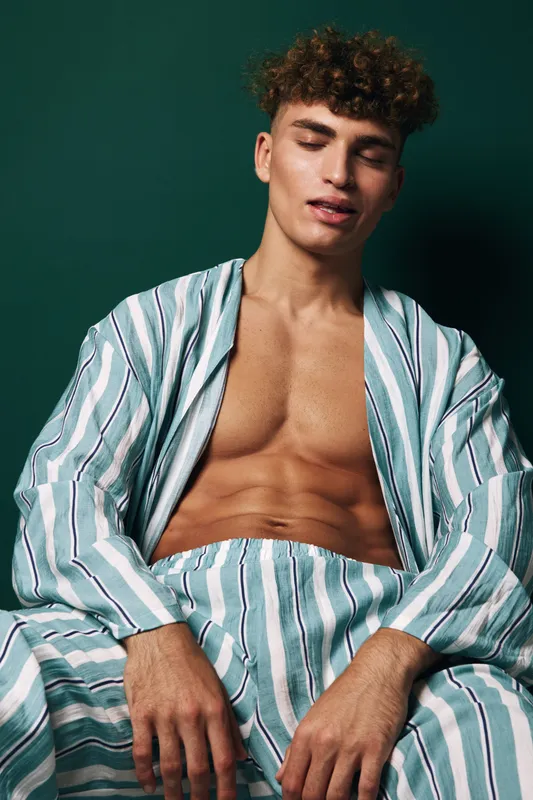 Young man with curly hair wearing striped pajamas sitting relaxed with closed eyes against dark green background. Casual style, comfortable clothing and peaceful expression.