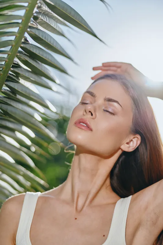 woman enjoying sunlight with closed eyes near tropical leaves, natural beauty, peaceful mood, summer vibes, clear skin glowing softly under bright sun