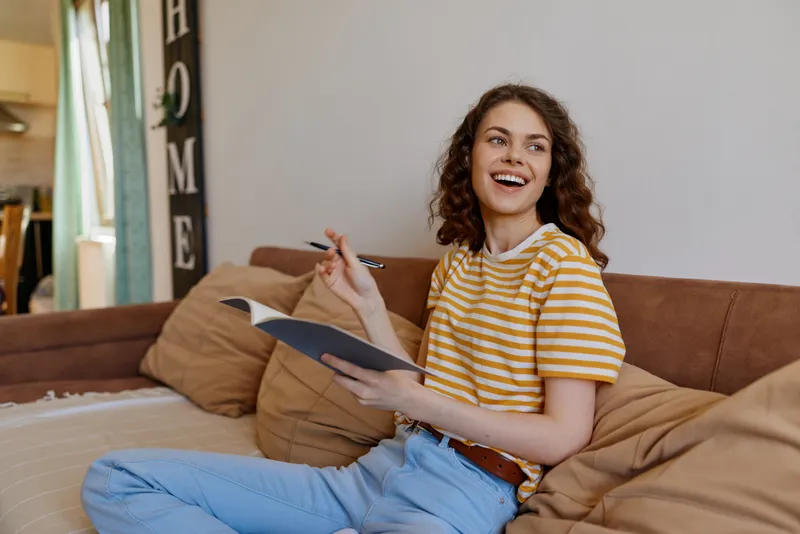 Young woman sits on a cozy couch at home, smiling while holding a notebook in a striped yellow and white tee. On a bright afternoon with natural light, creating a warm, inviting vibe.
