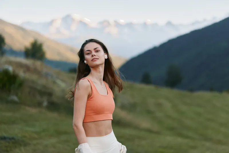 A confident young woman stands on a grassy hillside with distant mountains in the background. She wears an orange athletic top and light pants, gazing toward the camera.