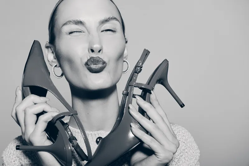 Smiling Caucasian woman with fair skin holding two high heels, puckering lips. Concept of fashion, style, and footwear accessories.