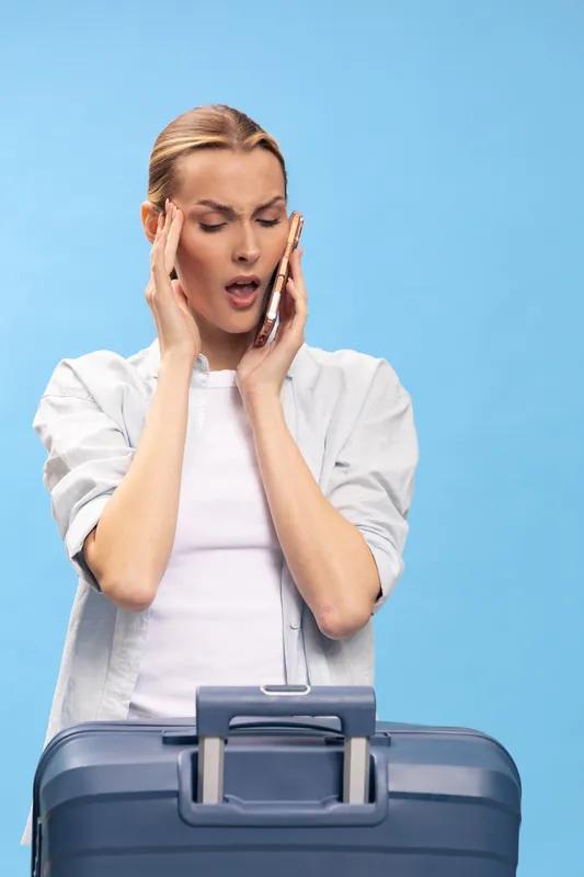 Stressed young woman in casual clothes holds phone to ear and touches temple, standing behind suitcase on blue background. Concept for travel problems, anxiety, and urgent communication.