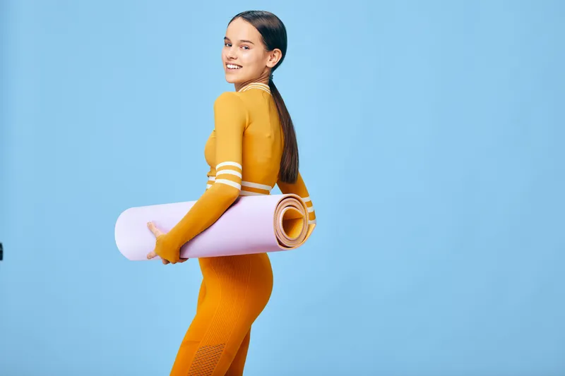 Woman in orange athletic wear carrying a rolled yoga mat, ready for a workout. Bright studio backdrop in blue highlights fitness, health and active lifestyle.