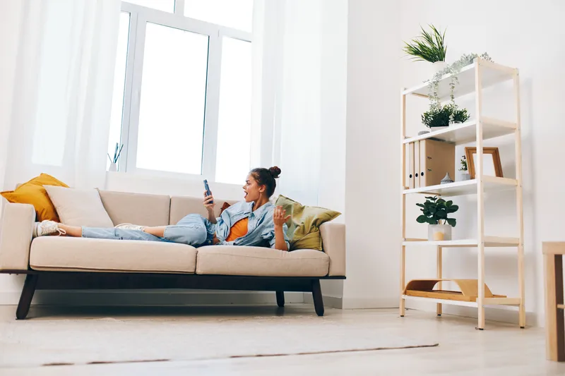 Счастливая женщина держит мобильный телефон и отдыхает на диване дома, погрузившись в досуг в киберпространстве
