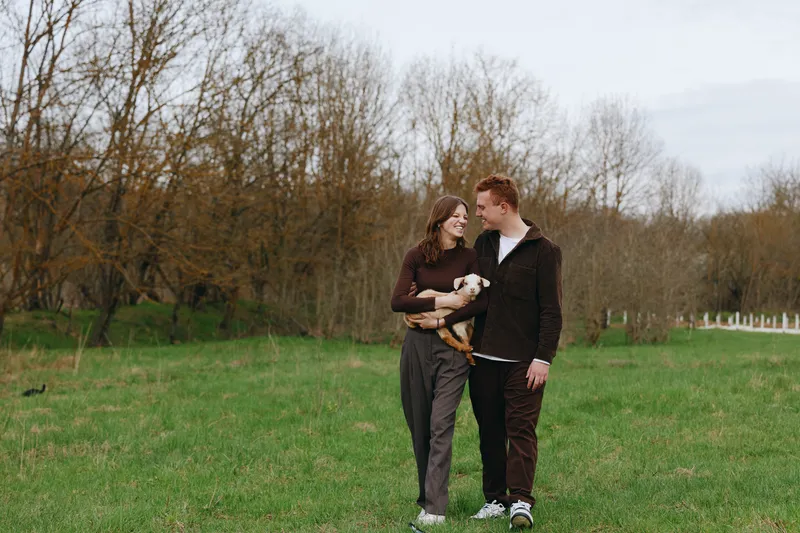 A smiling couple strolls across a sunny park, holding a tiny goat kid. A candid lifestyle moment of affection, companionship, and harmony with nature. A light, joyful moment. Outdoors.