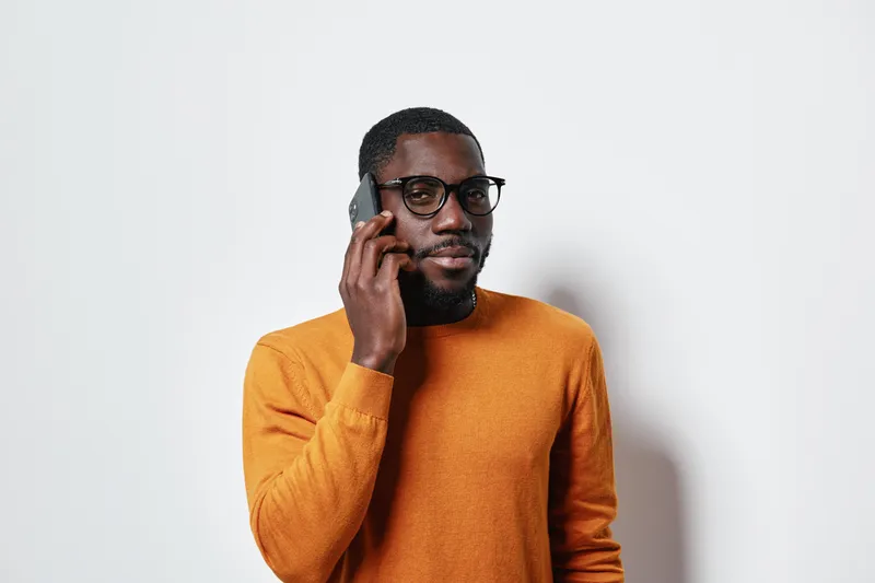 African American man with dark skin wearing glasses and orange sweater talking on smartphone against white background. Communication, technology and casual style with confident expression.