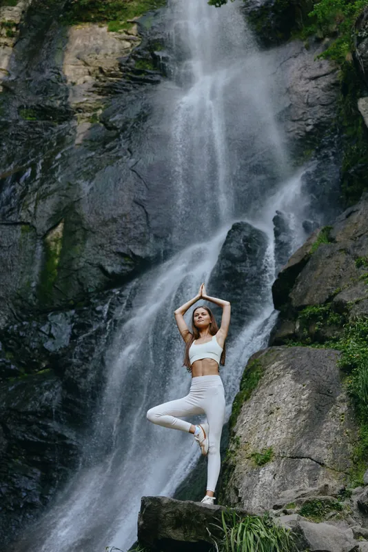 woman practicing yoga stands in tree pose near waterfall surrounded by rocks and lush greenery in serene nature setting during daytime