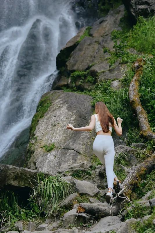 woman hiking near waterfall climbing rocky trail surrounded by lush green vegetation in natural outdoor setting