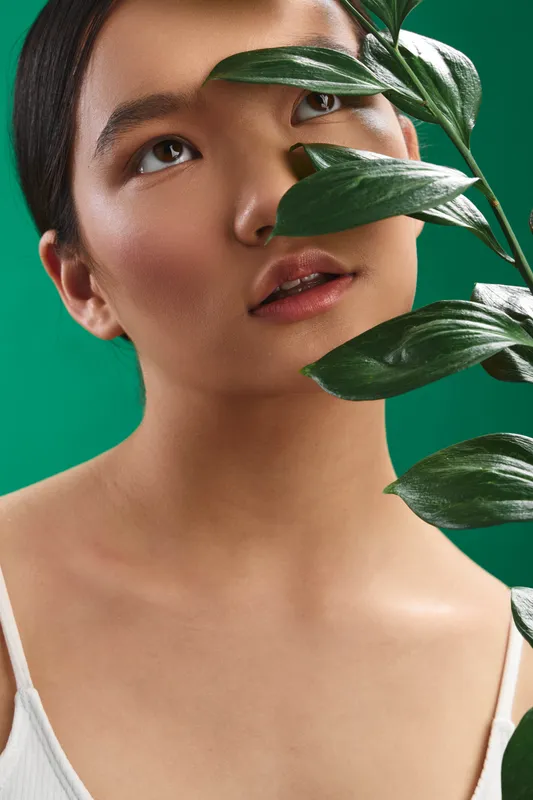Asian woman gazes softly beyond the frame, plant leaves cross her face, a vibrant green backdrop highlighting natural beauty and serene, fresh mood.