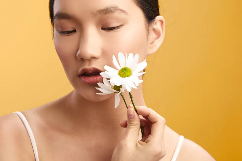 Asian woman holds a white daisy near her lips in a soft studio setting, calm expression, warm background, delicate beauty portrait conveying gentle mood and subtle elegance.