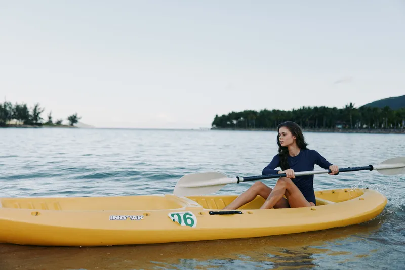 Person paddling a bright yellow kayak on calm water, wearing a dark shirt, oar in hand, shoreline with trees in the distance, conveying outdoor adventure and summer recreation.