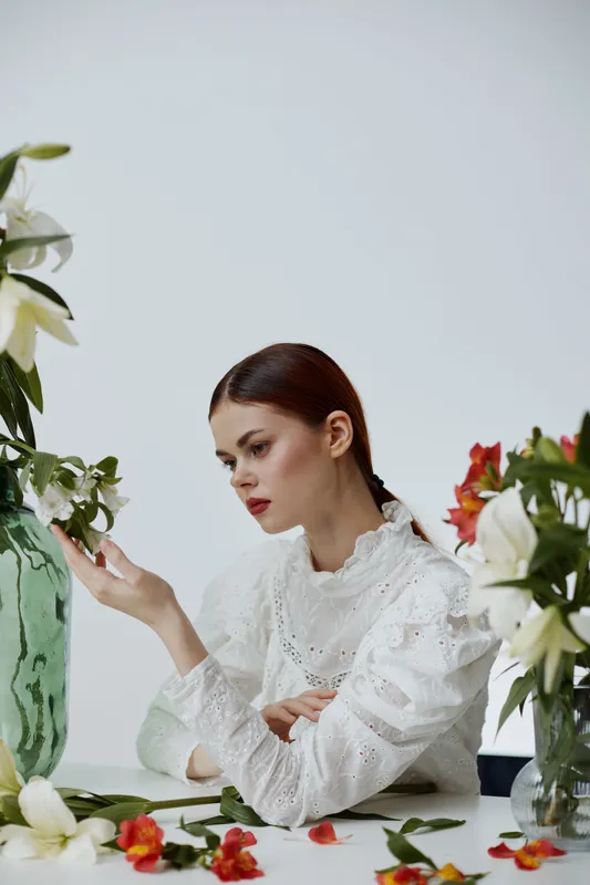 A young woman in a white blouse surrounded by vibrant flowers, reflecting on beauty and nature in a minimalistic setting