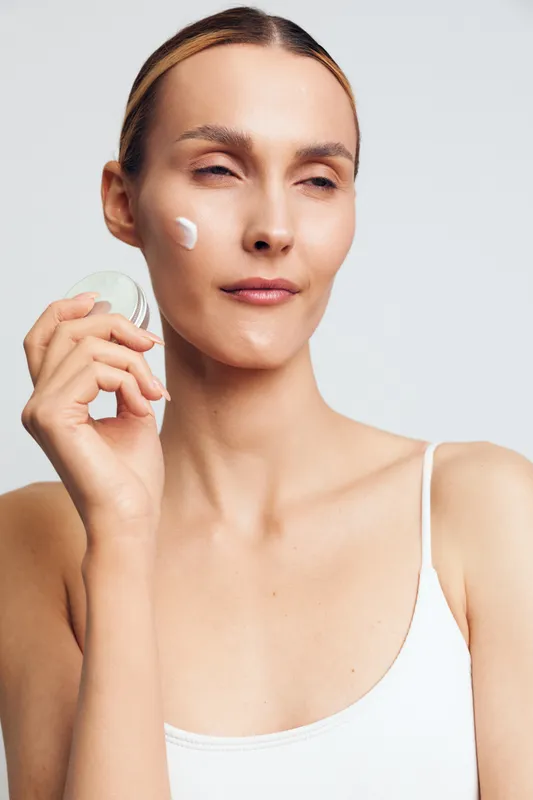 A woman with clear skin applies face cream using a cotton pad in a minimal white outfit against a light background. Concept for skincare, beauty routine, and self care.