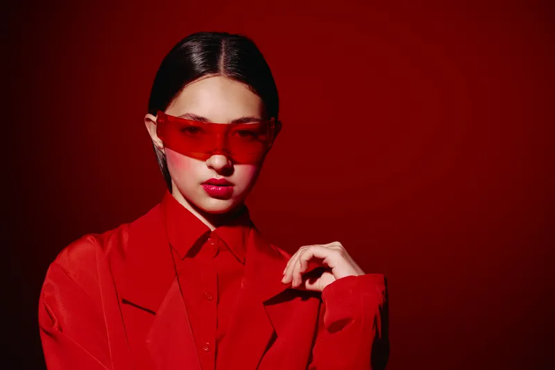 Young woman in red outfit and translucent red sunglasses poses against dark red background. High fashion beauty concept with monochrome intense style and confident expression.