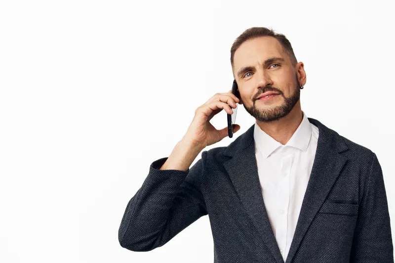 A professional business man on a clean white background, wearing a dark blazer and white shirt, speaks on a headset, projecting confident focus and friendly engagement.