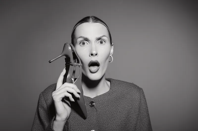 Surprised white woman with light skin holding high heel shoe near face, wearing casual clothing, isolated on grey background. Concept of shock, fashion and emotions.