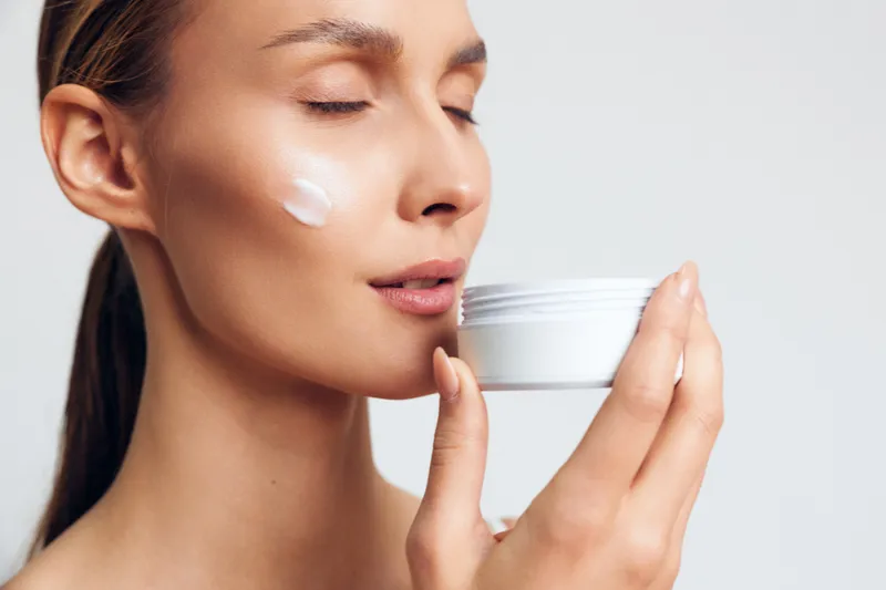 A calm young woman applies facial moisturizer in a minimal studio, showcasing a skincare routine and a small jar of cream in a close up moment of self care.