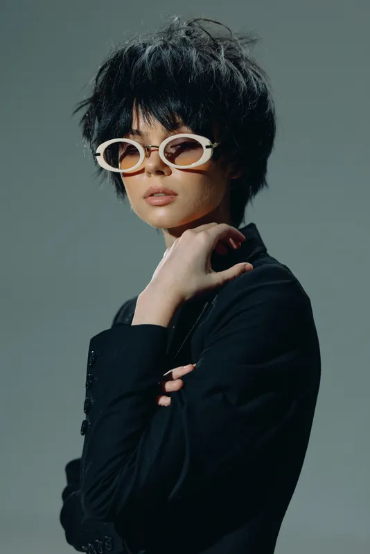 Stylish woman with short black hair and white sunglasses wearing a black outfit with a confident pose and neutral expression