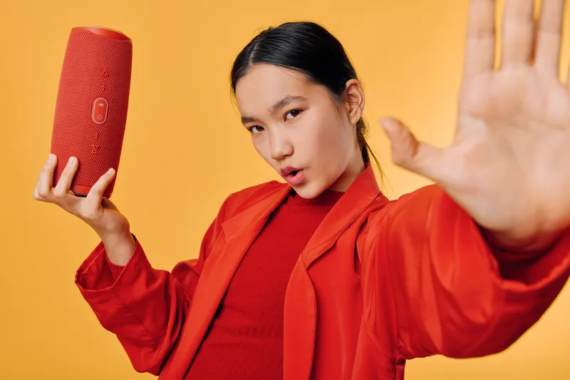 Woman in red outfit holding portable speaker with hand up on yellow background for technology music lifestyle and audio entertainment sound device.