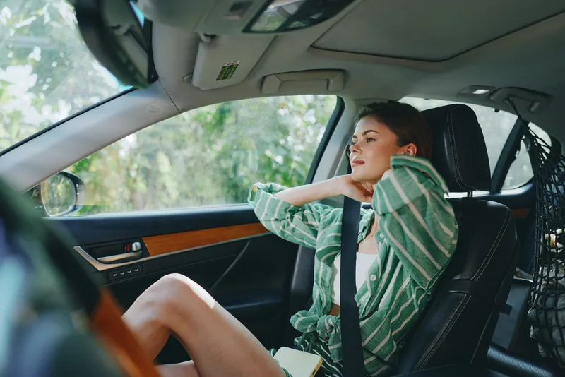 A relaxed woman sits in the driver's seat of a car, wearing a green striped shirt and a subtle smile, ready for a joyful journey.