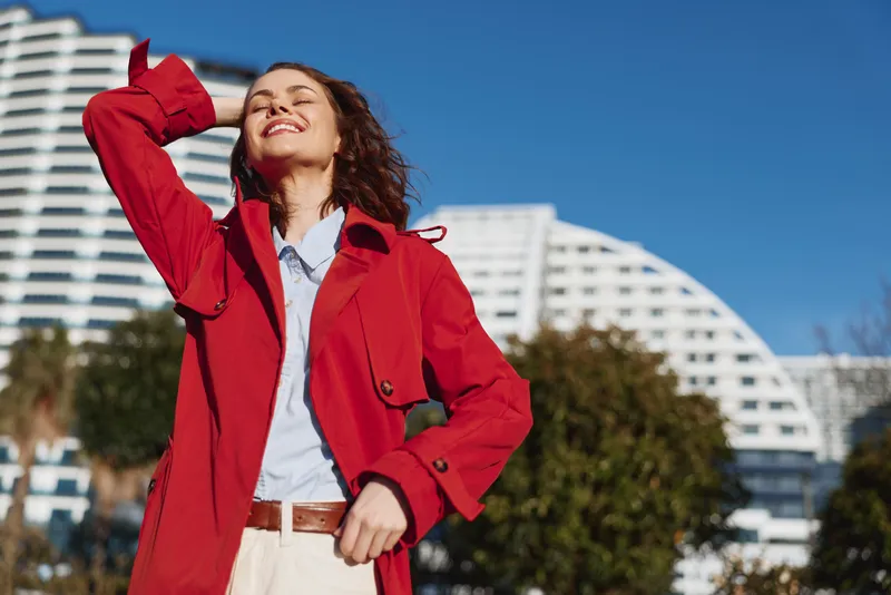 Young woman in a red coat enjoys a sunny day outdoors with eyes closed and a joyful smile, feeling relaxed and carefree in an urban environment.