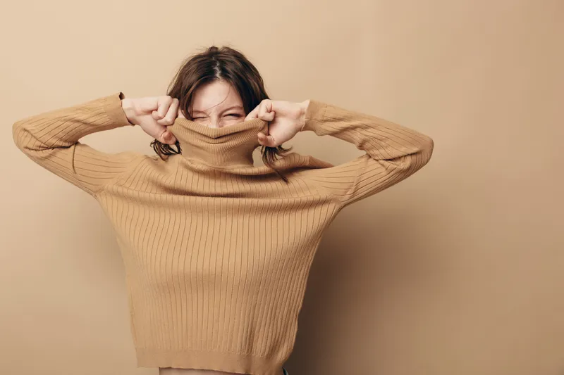 Young woman in beige sweater pulls collar over face with closed eyes, standing against matching background. Concept of cozy fashion, autumn style, and playful mood in studio.