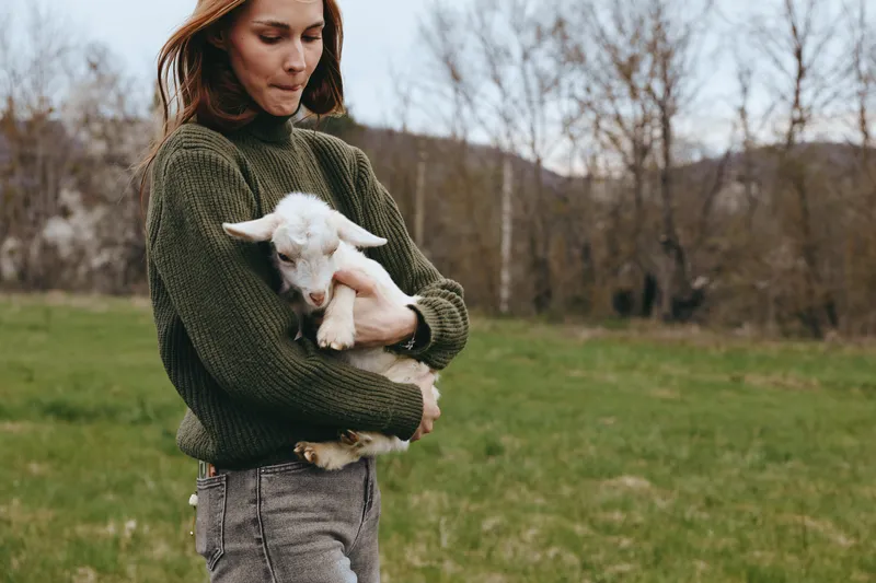 Candid woman outdoors, gently cradling a young goat kid in a cozy sweater, soft sunlight on a quiet rural field, relaxed posture, natural colors, wholesome farm moment, authentic lifestyle