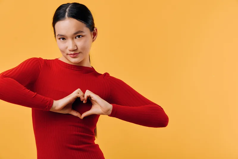 Young woman in red sweater making heart shape with hands against orange background. Casual gesture, studio portrait with smiling expression, love concept, isolated color wall, positive emotion.