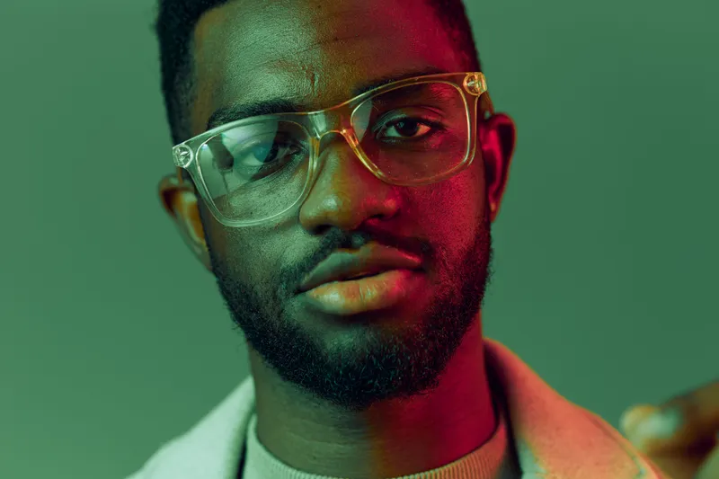 Portrait of a young Black man with glasses, expressing confidence against a vibrant green and pink background, ideal for concepts of individuality and self esteem