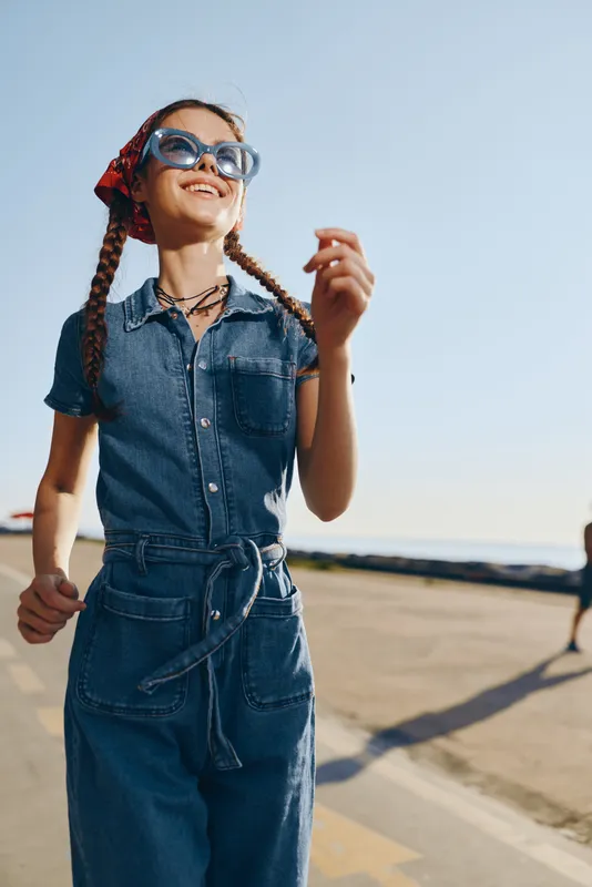 smiling woman in denim jumpsuit and sunglasses enjoying sunny day outdoors with casual style and braided hair in a bright clear sky setting