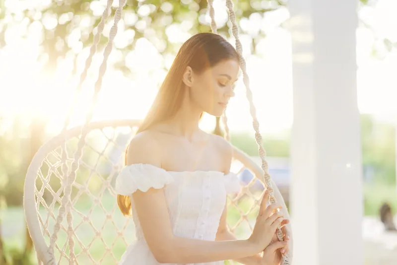 A serene woman in a white summer dress sitting on a swing, gazing thoughtfully, surrounded by lush greenery and soft sunlight, evoking feelings of peace and contemplation