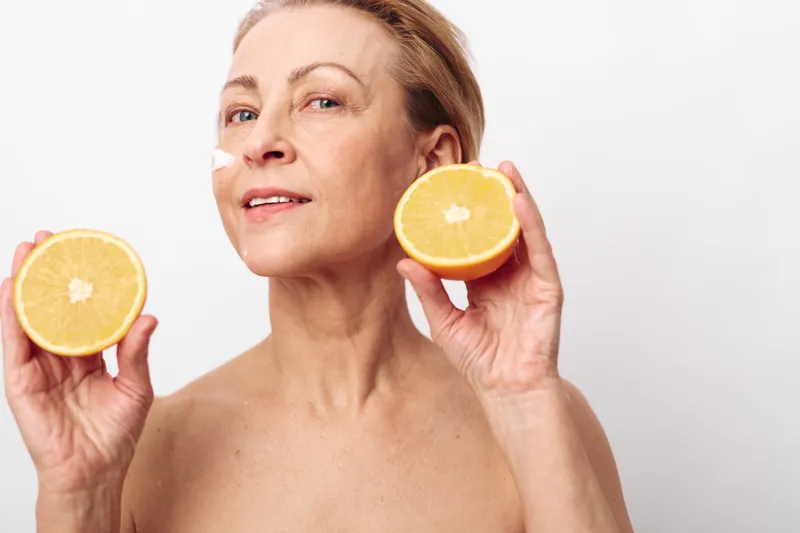 Mature woman holds two halves of a fresh orange near her face, showing natural skin and healthy lifestyle. White background highlights wellness and citrus fruit concept.