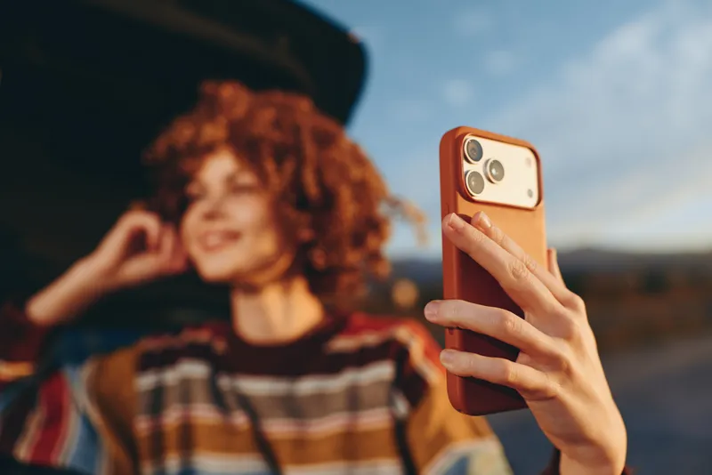 Woman lifestyle selfie, hand holds smartphone, curly haired woman smiles in soft focus outdoors on urban street, striped sweater and hat, warm golden travel light