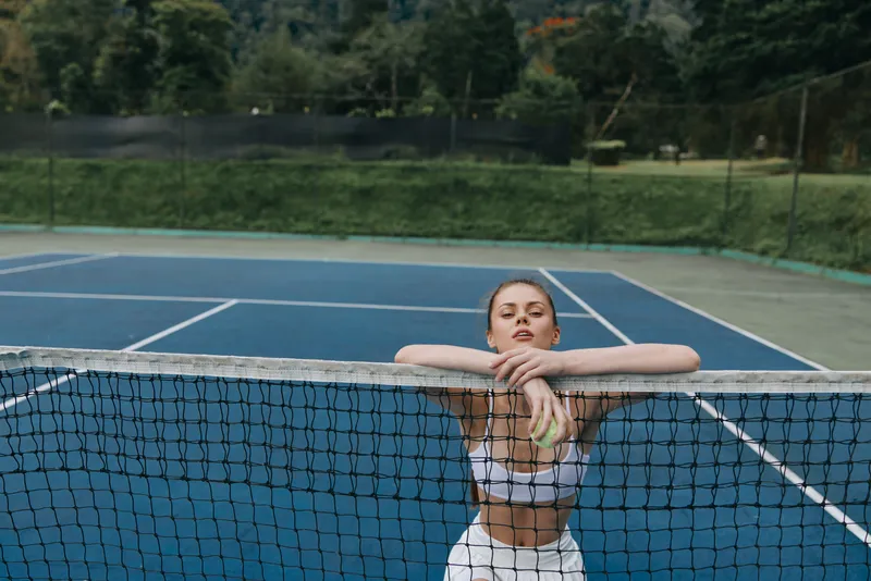 Sporty young woman resting on tennis net, wearing stylish sportswear, exuding confidence, set against lush green nature in a vibrant blue tennis court