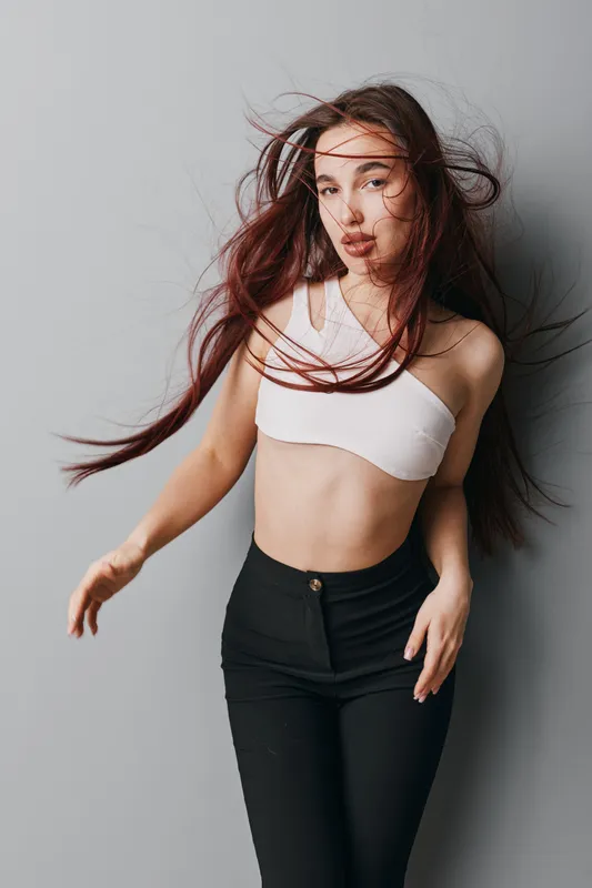 Young woman with long hair wearing white crop top and black pants posing against gray wall with hair flowing in motion, fashion style and confident expression in studio lighting.