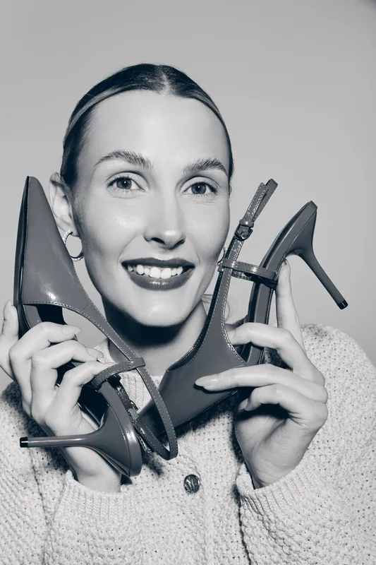 Happy white woman with fair skin holds high heel shoes near face smiling, wears knitted sweater. Concept of fashion accessories, footwear, and positive lifestyle.