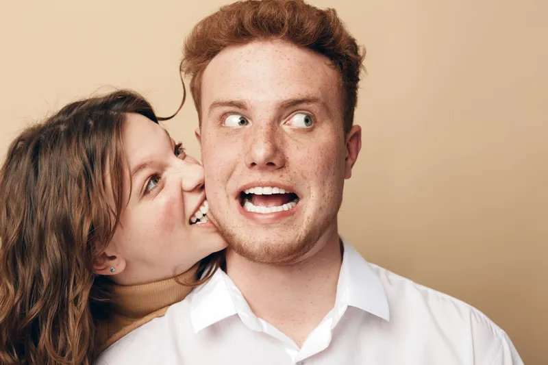 Playful young woman bites cheek of surprised redhead man in white shirt. Studio portrait on beige background. Concept of fun, friendship, humor, and candid emotions between couple.