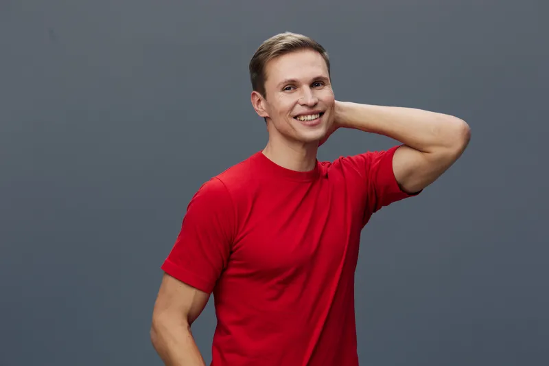 Bright cheerful young man with short light brown hair wearing casual red t-shirt smiling and looking directly at camera while standing against plain blue background. People lifestyle concept, happines