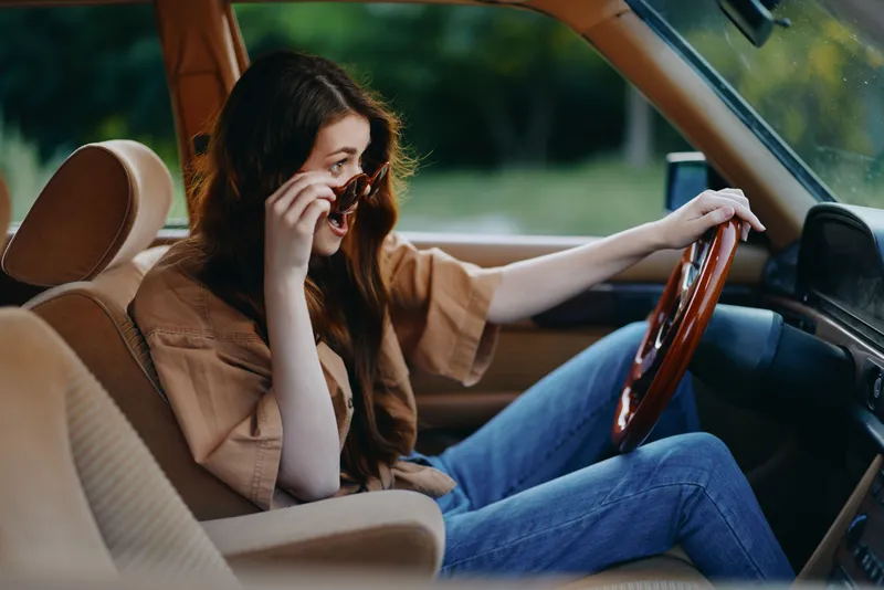 Young woman in casual outfit driving a vintage car, smiling and adjusting sunglasses, enjoying a sunny day outdoors, perfect for lifestyle and travel concepts.