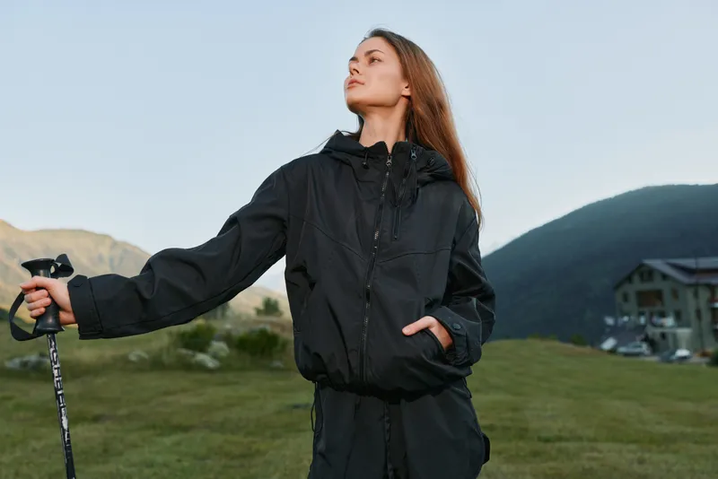 A cheerful hiker stands outdoors on a windy mountain landscape, wearing a black windbreaker and gripping a trekking pole. She gazes upward with confidence, signaling adventure and energy.