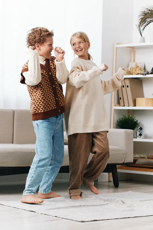 Two young children dance joyfully indoors on a bright living room rug, wearing cozy sweaters and jeans, capturing candid happiness and playful friendship.