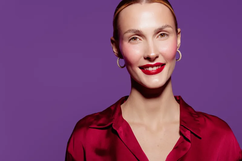 Smiling woman with red lipstick and earrings wears red satin blouse on purple background. Portrait of confident female model with natural skin and bright makeup. Studio fashion concept.