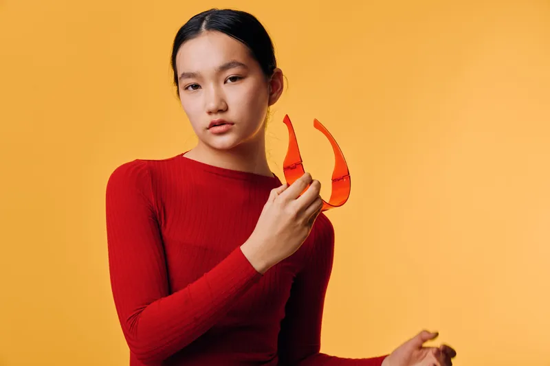 Young woman in red sweater holding orange lobster claws with serious expression on yellow background. Portrait of Asian female with minimal makeup and casual pose indoors.