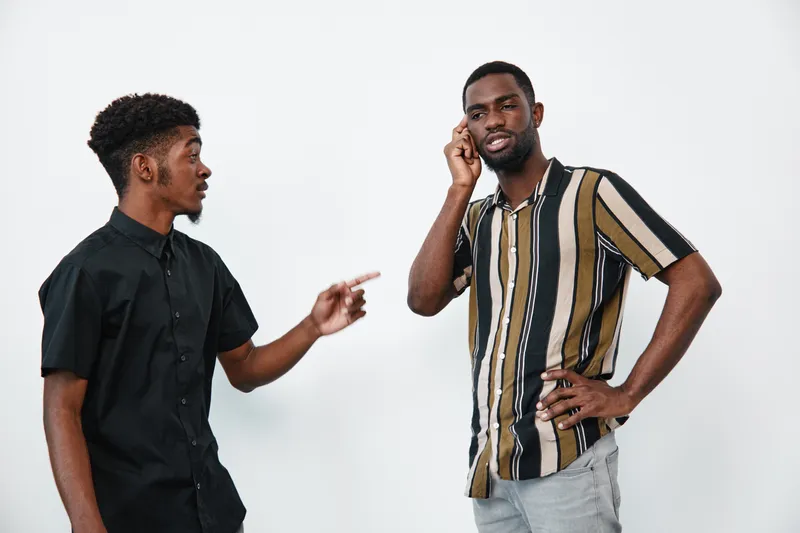 Two African American men with dark skin having a conversation on white background. One man is pointing while the other listens with hand on chin in casual clothes.