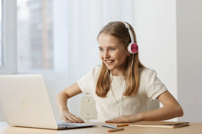 Happy young girl with headphones engaging in online learning, smiling at a laptop in a bright room, showcasing modern education and enthusiasm for learning