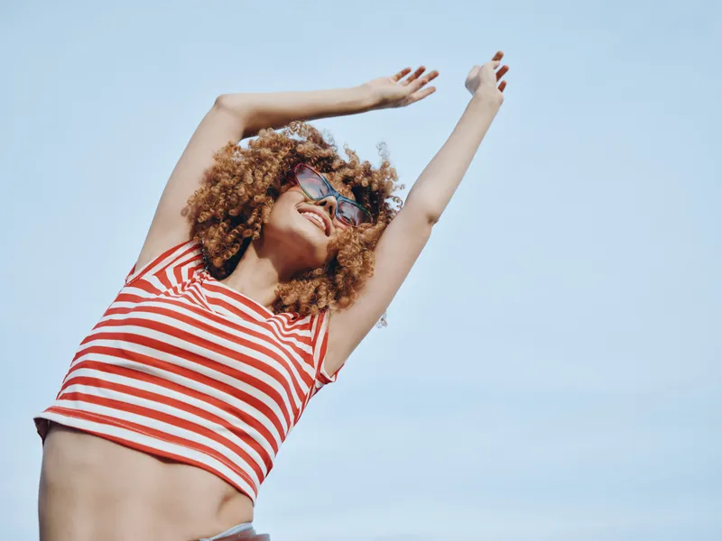 Vibrant portrait of a joyful woman posing outdoors under a clear blue sky, wearing a red and white striped top and sunglasses, arms raised in a carefree summer moment of celebration.